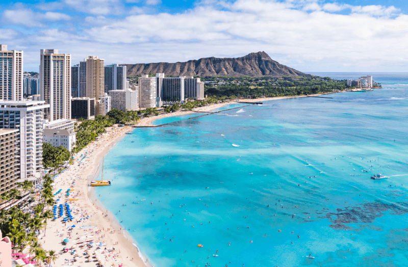 Aerial view of coast surrounded by the sea and buildings, with mountains and clear sky in the background