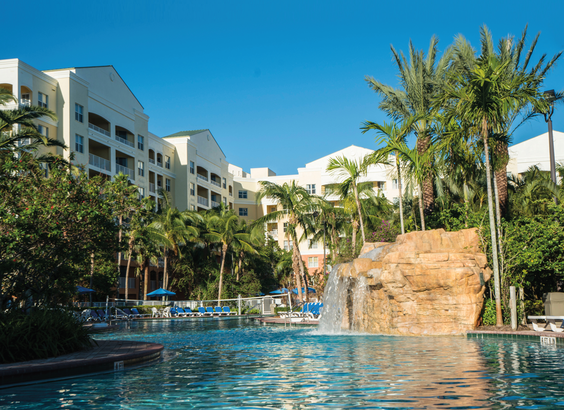 View of a swimming pool with artificial waterfall surrounded by palm trees and buildings, with clear sky in the background