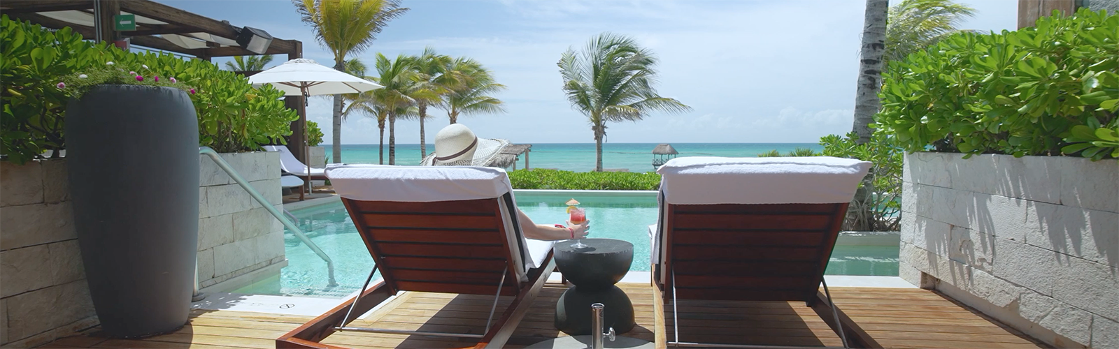 View from behind of a person lying on a lounge chair and holding a drink in front of a swimming pool, with palm trees and the sea in the background