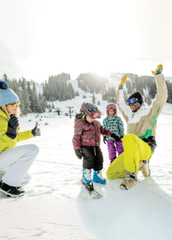 Two adults and two children playing in the snow, with trees and snowy mountains in the background